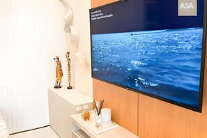 Una habitación con un televisor de pantalla plana grande que muestra una vista al mar, un mueble de madera con un florero blanco y bolígrafos, y una escultura sobre un estante blanco.