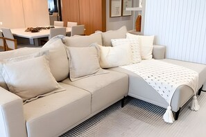 A modern living room with a beige sectional sofa, a patterned throw blanket, and a dining area with white chairs and a table.