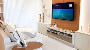 A modern living room with a flat-screen TV mounted on a wooden wall, a white sofa, and a small coffee table. - ASA Apto Vale Dourado Frente-mar Hidro 2vgs (Balneário Camboriú)
