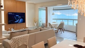 A modern living room with a large flat-screen TV, a sofa, a dining table, and a balcony with a view. - ASA Apto Vale Dourado Frente-mar Hidro 2vgs (Balneário Camboriú)