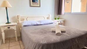 A neatly made bed with a striped blanket and two pillows, a bedside table with a lamp, and a small bedside table with a green object on it. - ASA Frente Mar Pe na Areia 4QT BC (Balneário Camboriú)