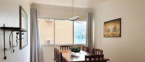 A dining area with a table, chairs, a window with a city view, a painting on the wall, and a hanging light fixture.