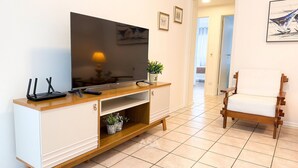 A living room with a flat-screen TV, a wooden entertainment unit, a white armchair, and a tiled floor. - ASA Frente Mar Pe na Areia 4QT BC (Balneário Camboriú)