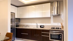 A modern kitchen with white cabinets, a stainless steel range hood, a built-in oven, and a microwave. - ASA Apto Praia dos Amores 2 Suites BC (Balneário Camboriú)