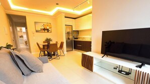 Ein modernes Wohnzimmer mit Flachbildfernseher, einer Couch, einem Essbereich mit Stühlen und einer Küche inklusive Kühlschrank und Mikrowelle.