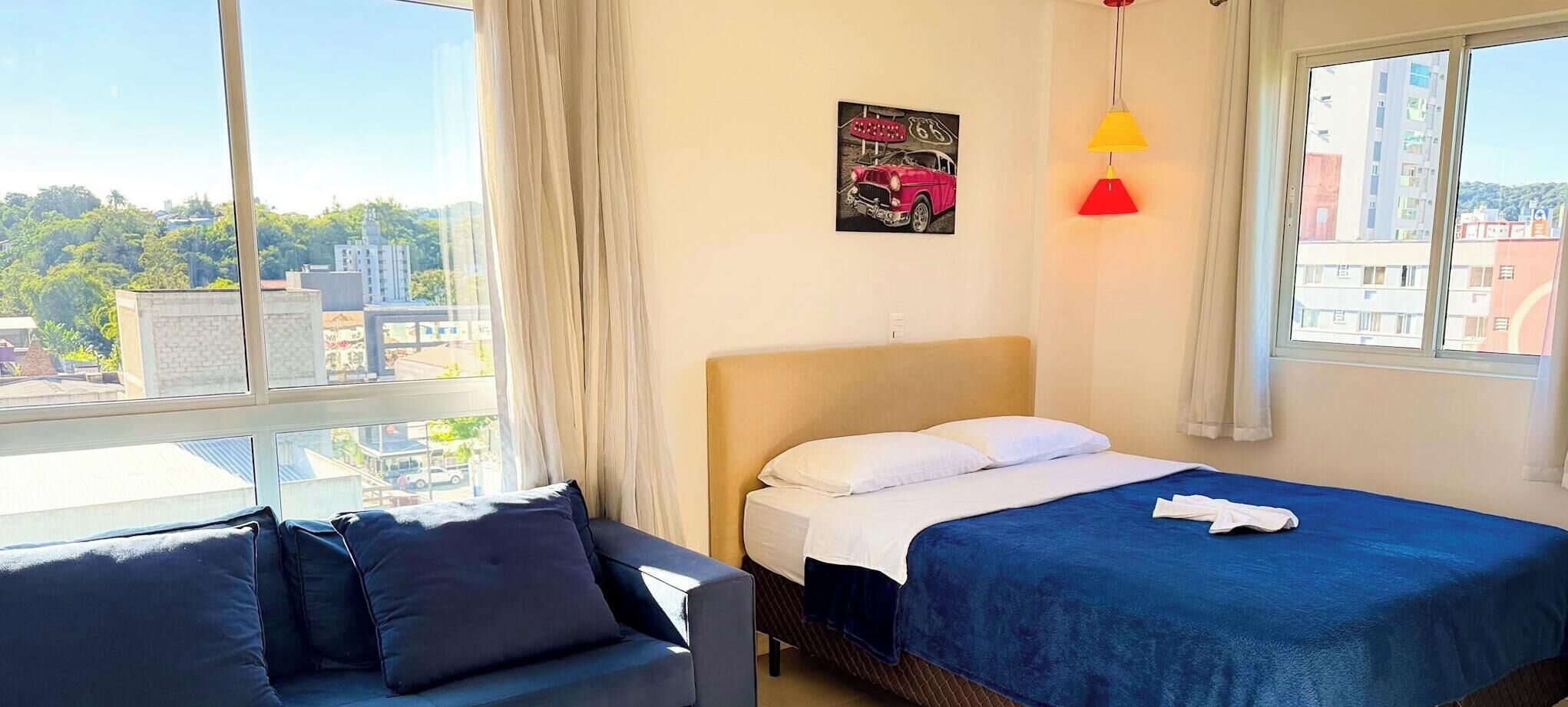 A hotel room with a bed, a sofa, a window with a city view, and a painting on the wall.