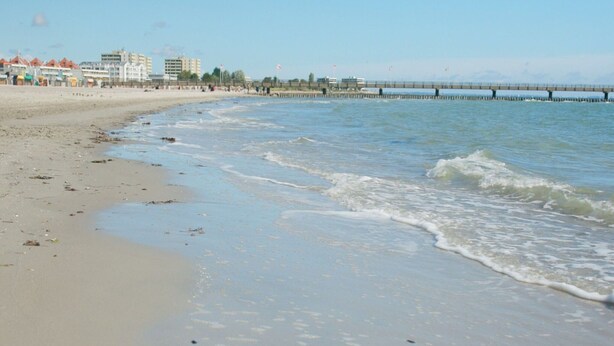 A beach with a pier extending into the water, buildings in the background, and a clear sky. - Geraumiges Wellness-zuhause fur 6 Personen (Grossenbrode)