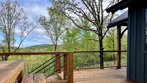 Una terraza de madera con una escalera que conduce a un área cubierta, rodeada de árboles y vistas del paisaje.
