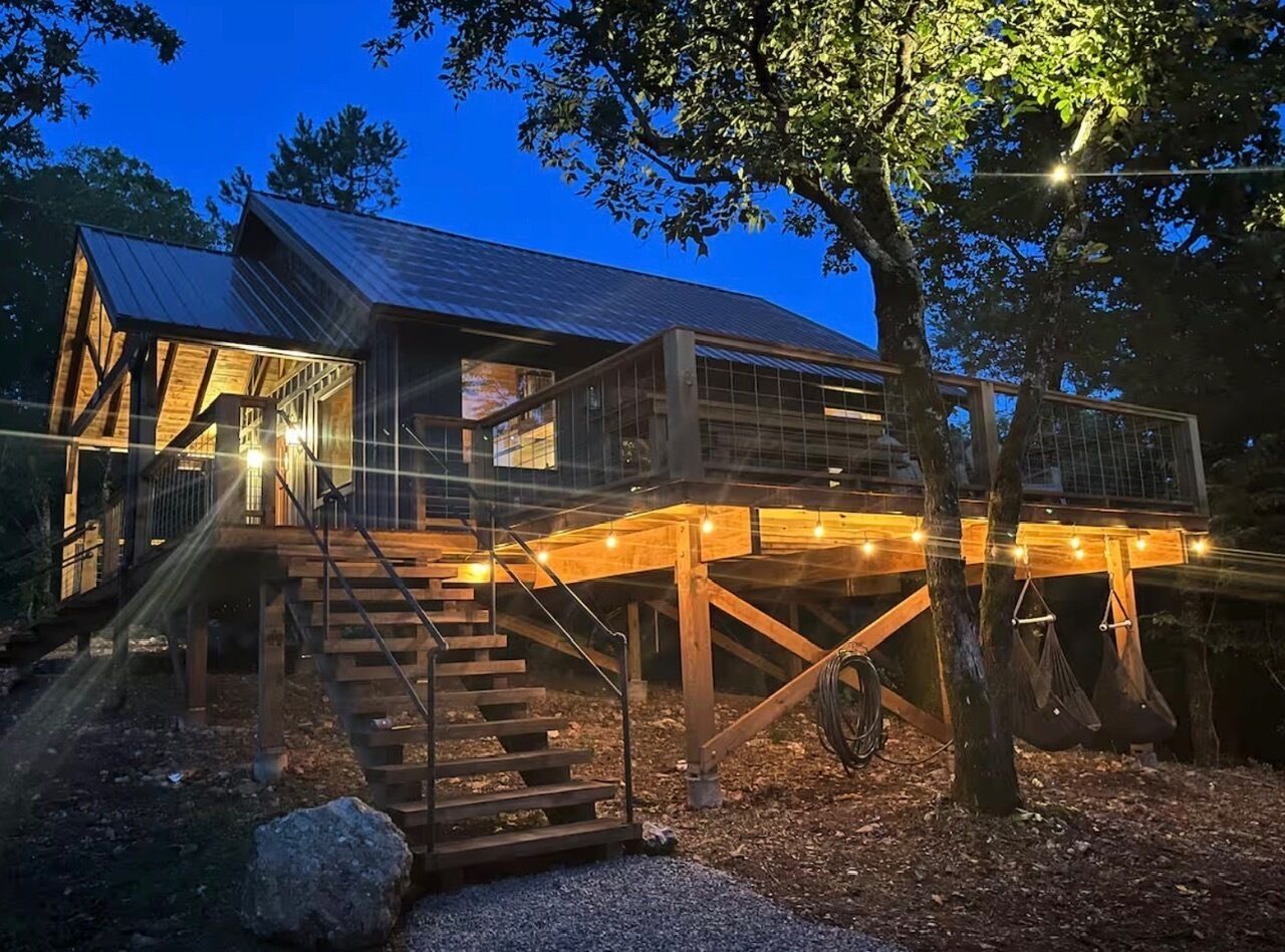 A two-story wooden cabin with a balcony, wooden stairs, and string lights.