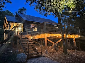 A two-story wooden cabin with a balcony, wooden stairs, and string lights.