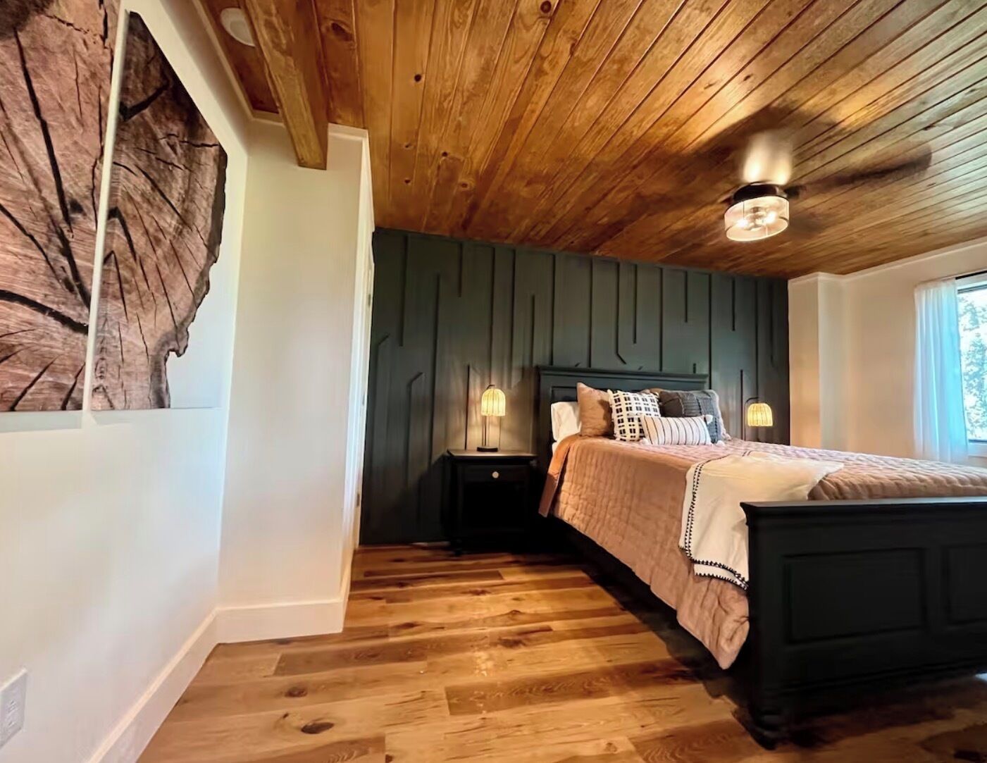 Une chambre avec un plafond en bois, un lit doté d’une tête de lit sombre, deux lampes de chevet et une grande œuvre d’art murale en bois.