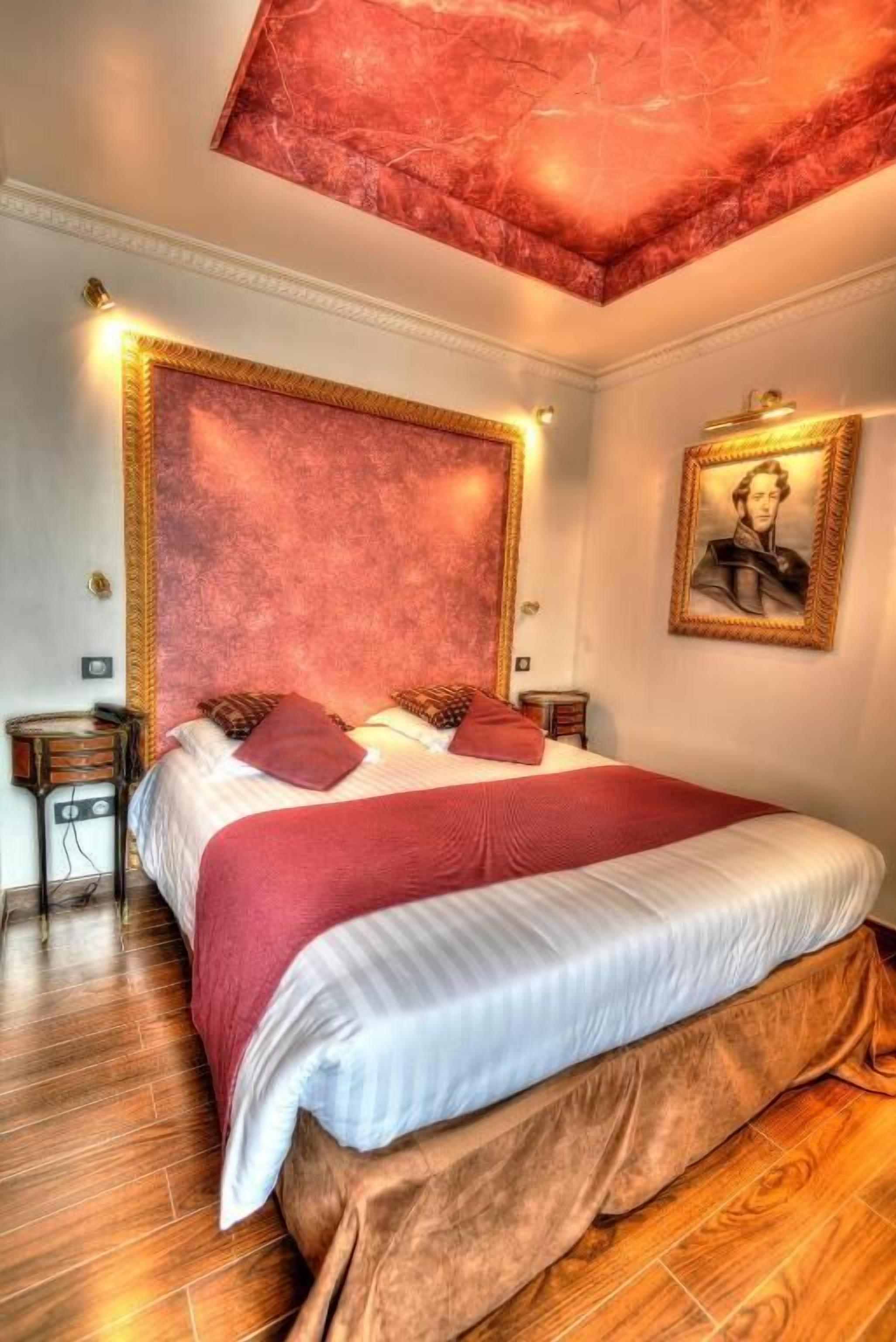 Een hotelkamer met een groot bed, een rode accentmuur, een lijst met een portret en een houten vloer.