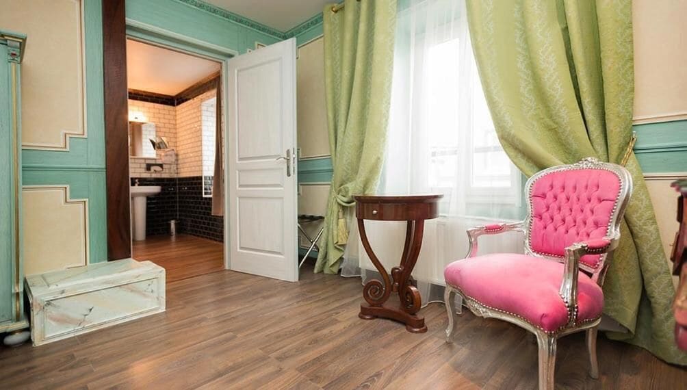 Een kamer met een houten vloer, een roze fauteuil, een klein houten tafeltje en een deur naar een badkamer.