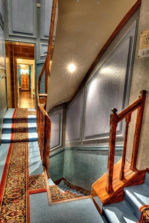 Un magnifique escalier orné de balustrades travaillées et recouvert d’un tapis à motifs, menant à une pièce dotée d’un grand miroir et d’une porte portant l’inscription « 4-9 ».