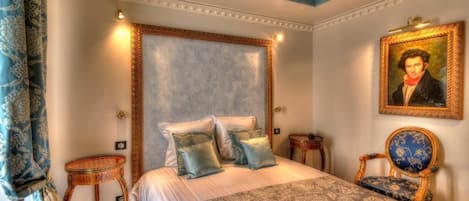 A bedroom with a large bed, two bedside tables, a chair, a painting on the wall, and a patterned bedspread.
