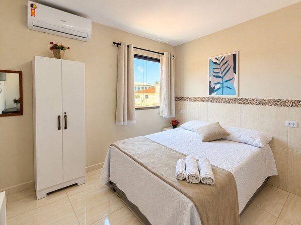 Une chambre d’hôtel comprenant un lit, une armoire, un appareil de climatisation, une fenêtre avec des rideaux et une œuvre d’art accrochée au mur.