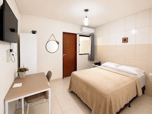 Habitación de hotel con cama, mesita de noche, silla, escritorio, televisor, aire acondicionado y ventana con cortinas.