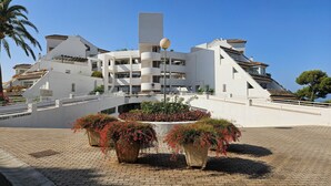 A modern building with a central tower, multiple balconies, and a paved area with flower planters. - Beautiful apartment with pool & sea views (Mijas Costa)