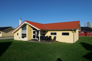 Ein einstöckiges Haus mit rotem Dach, einer überdachten Terrasse mit Gartenmöbeln und einem gepflegten Rasen.