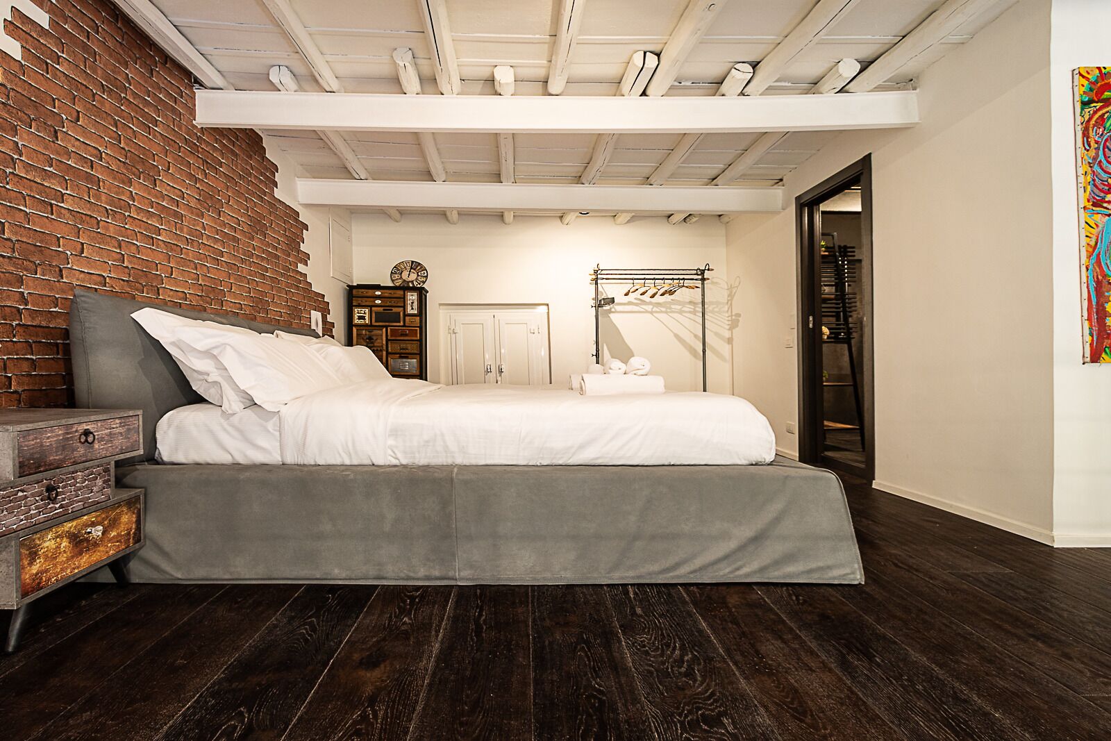 A bedroom with a brick wall, a bed, a nightstand, a clock, and a door leading to another room.
