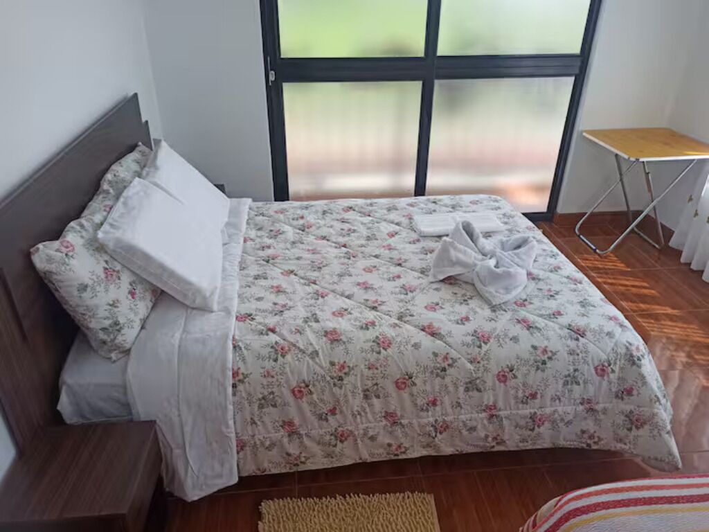 Un lit bien fait, recouvert d’une couvre-lit à motifs floraux et agrémenté d’oreillers blancs, une petite table de chevet en bois et une fenêtre donnant sur un paysage verdoyant.