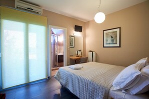 Une chambre d’hôtel comprenant un lit, un bureau avec une chaise, un radiateur, un tableau encadré au mur et une porte coulissante donnant accès à une salle de bain.