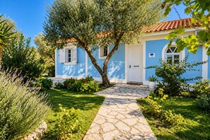 A house with a stone pathway leading to the entrance, surrounded by lush greenery and a clear blue sky. - Villa Thalia Galini - Two Bedroom Villa, Sleeps 4 (Vassilikos)