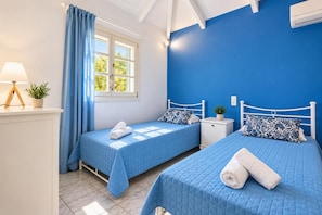 A room with two beds, a window with curtains, a bedside table with a lamp, and a potted plant. - Villa Thalia Galini - Two Bedroom Villa, Sleeps 4 (Vassilikos)