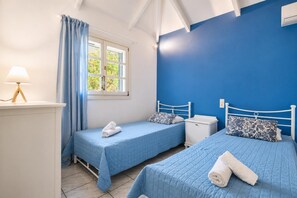 A room with two beds, a window with curtains, a bedside table with a lamp, and a wall painted blue. - Villa Thalia Galini - Two Bedroom Villa, Sleeps 4 (Vassilikos)