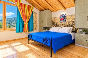 A bedroom with a bed, wooden flooring, a stone wall, a window with curtains, and a view of the landscape. - Villa Tireda Tria - Three Bedroom Villa, Sleeps 6 (Koridallos)