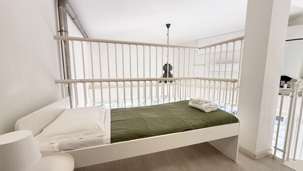 Ein helles Zimmer mit einem Einzelbett, einer grünen Tagesdecke, weißen Kissen und einem weißen Geländer.