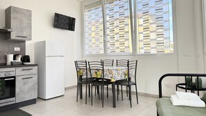 Eine kompakte Küche mit Kühlschrank, Herd und Mikrowelle. Ein Esstisch mit Stühlen sowie ein Bett mit grüner Bettdecke sind vorhanden.