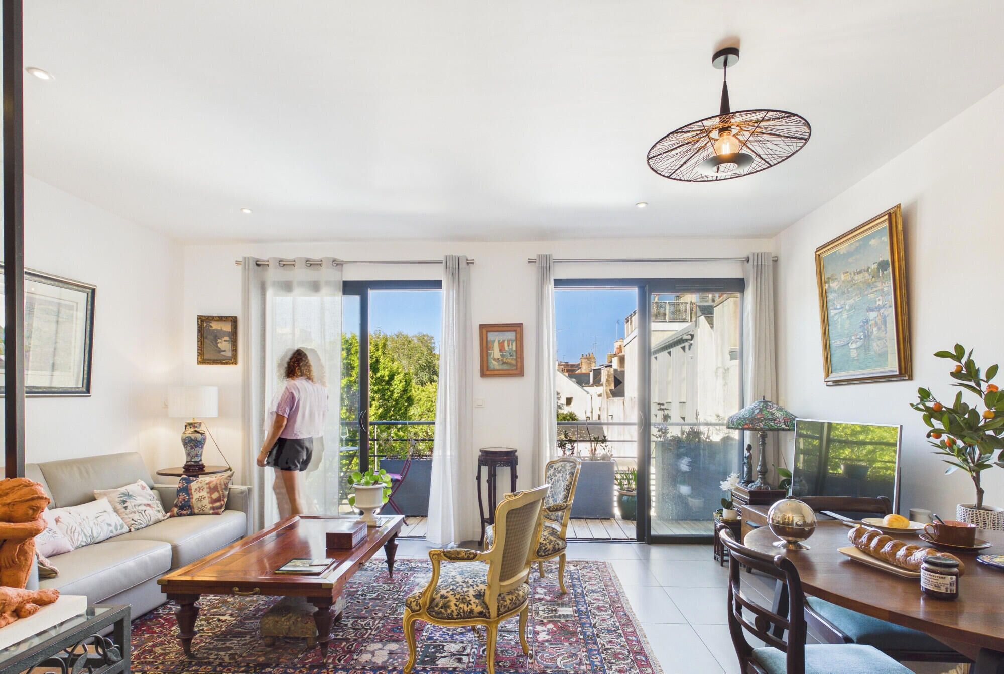 Un salon spacieux avec un canapé, une table basse et des fauteuils, un coin repas avec une table et des chaises, et un balcon offrant une vue sur les immeubles et les arbres.