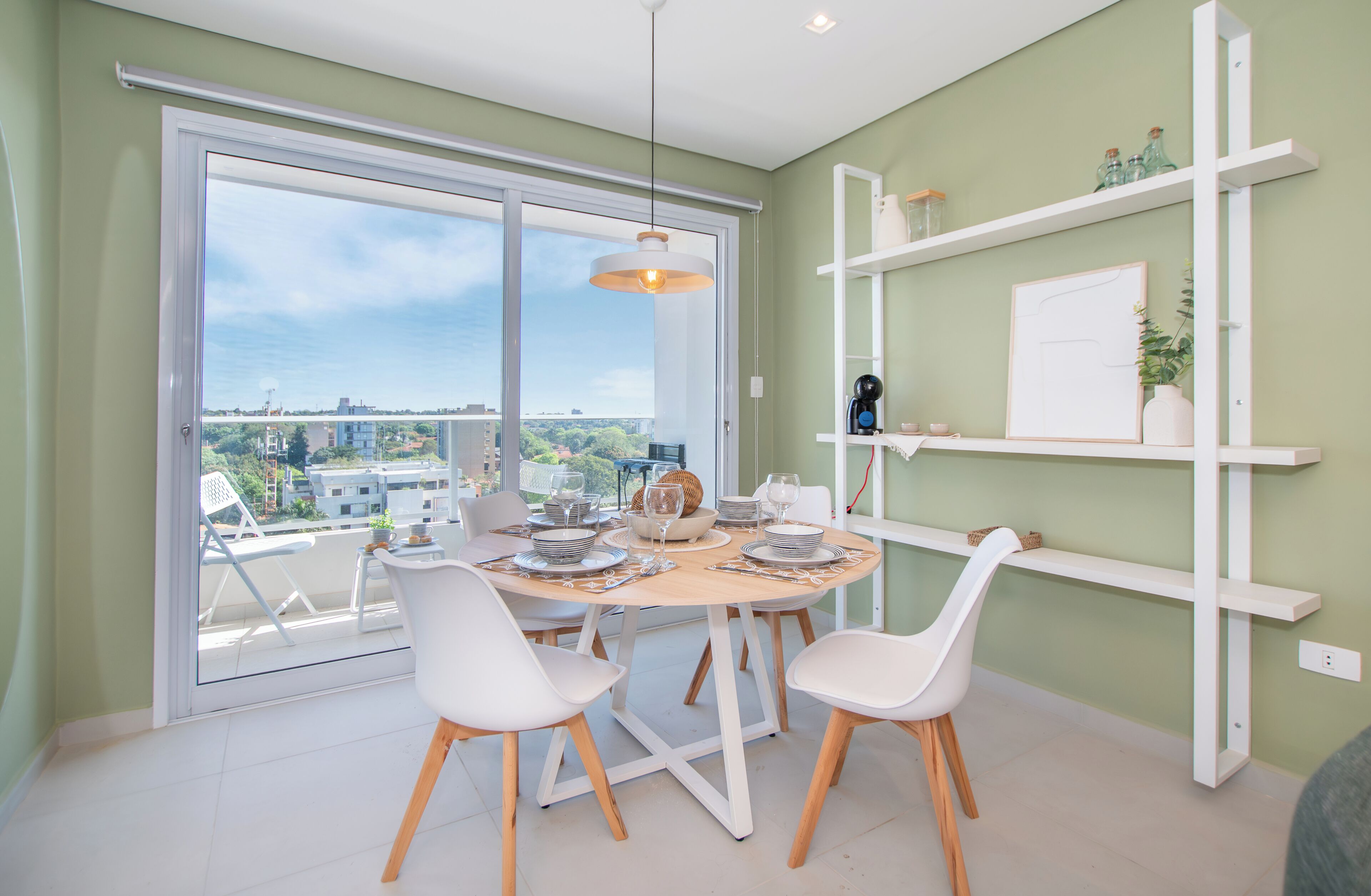A dining area with a table set for four, white chairs, and a view of the city through large windows.