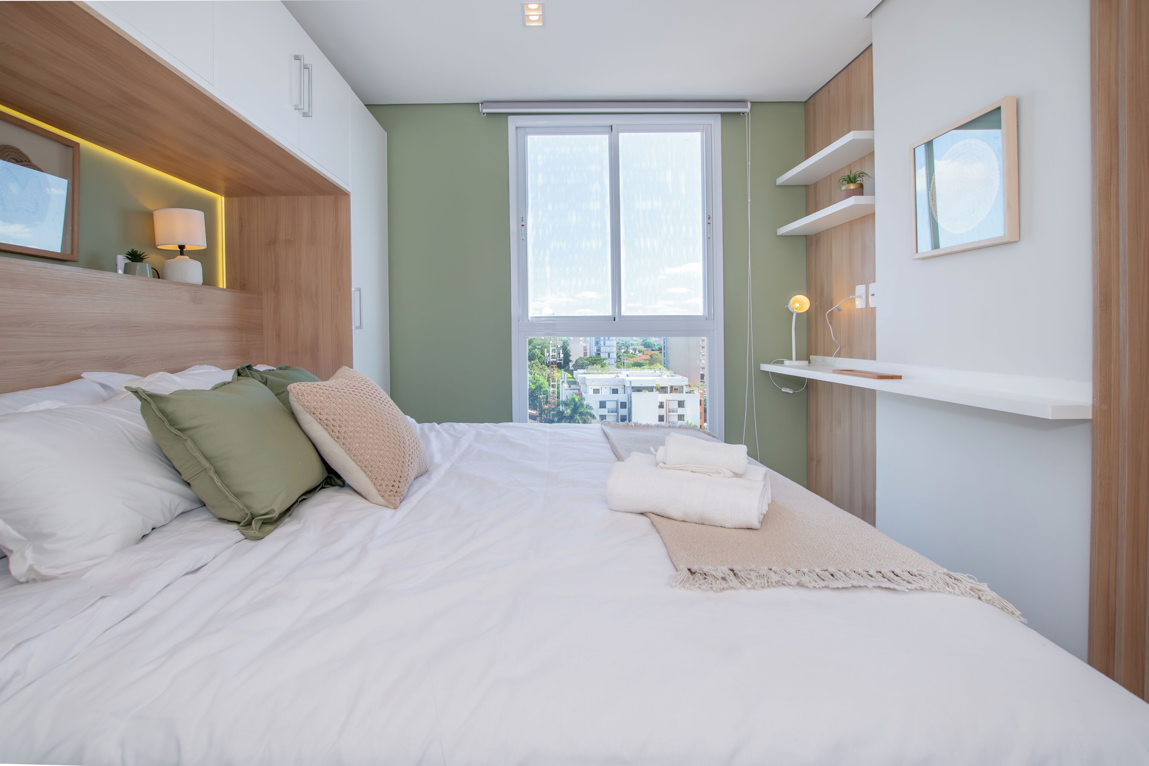 Een moderne slaapkamer met een groot bed, uitzicht op gebouwen door het raam en een plank met een lamp.