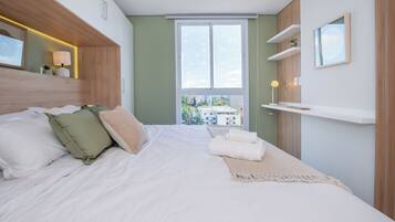 Een moderne slaapkamer met een groot bed, uitzicht op gebouwen door het raam en een plank met een lamp.