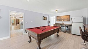 A pool table in a room with a kitchen area, a dining table, and a refrigerator.