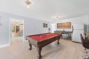 A pool table in a room with a kitchen area, a dining table, and a refrigerator.