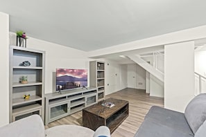 A modern living room with a flat-screen TV, wooden coffee table, and grey sofas.