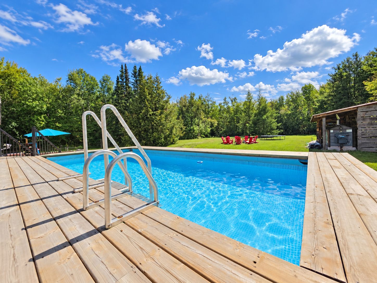 Ein Schwimmbecken mit Holzterrasse, umgeben von Bäumen und blauem Himmel.