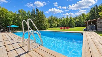 Ein Schwimmbecken mit Holzterrasse, umgeben von Bäumen und blauem Himmel.