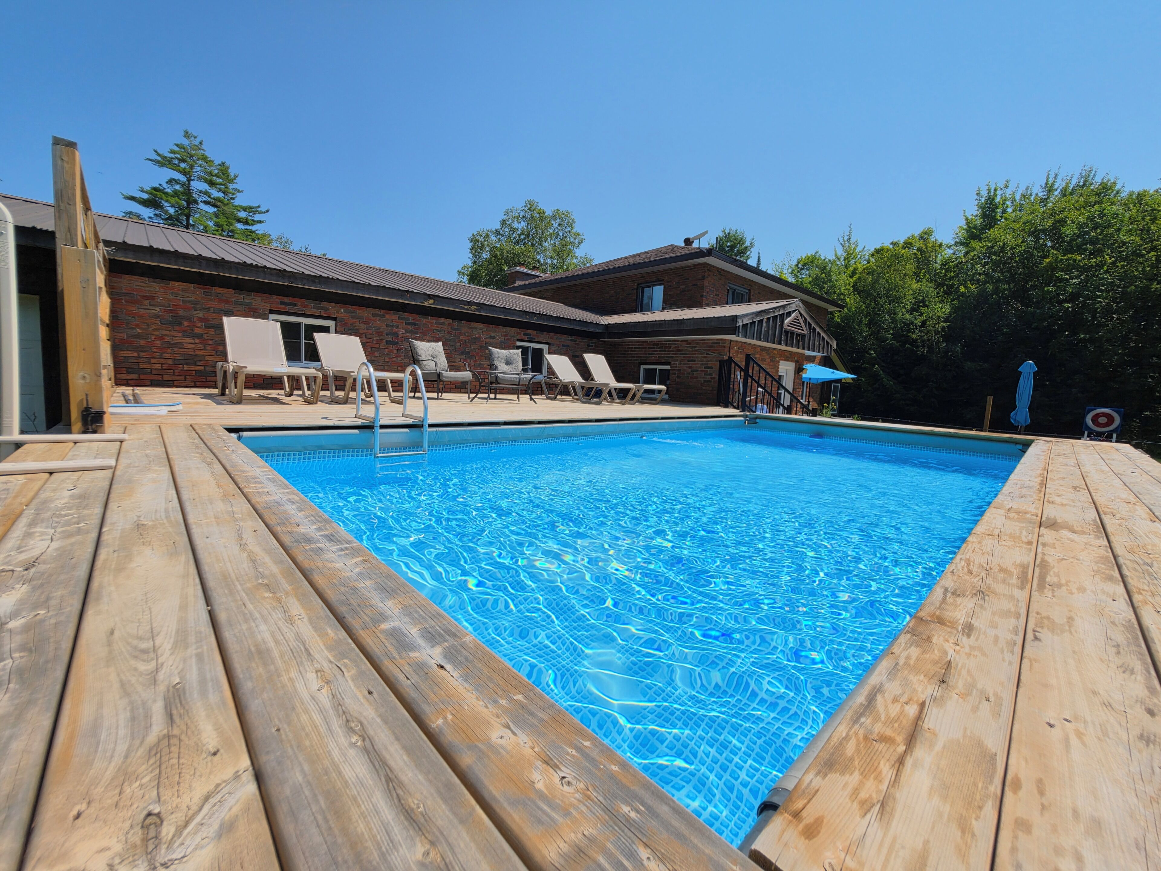 Una terraza de madera rodea una piscina con sillones de descanso y un edificio de ladrillo al fondo.
