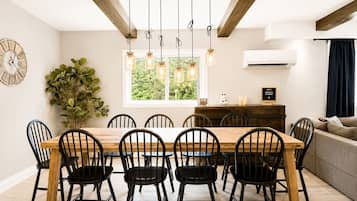 A dining area with a wooden table and black chairs, a large potted plant, and a view of greenery outside.