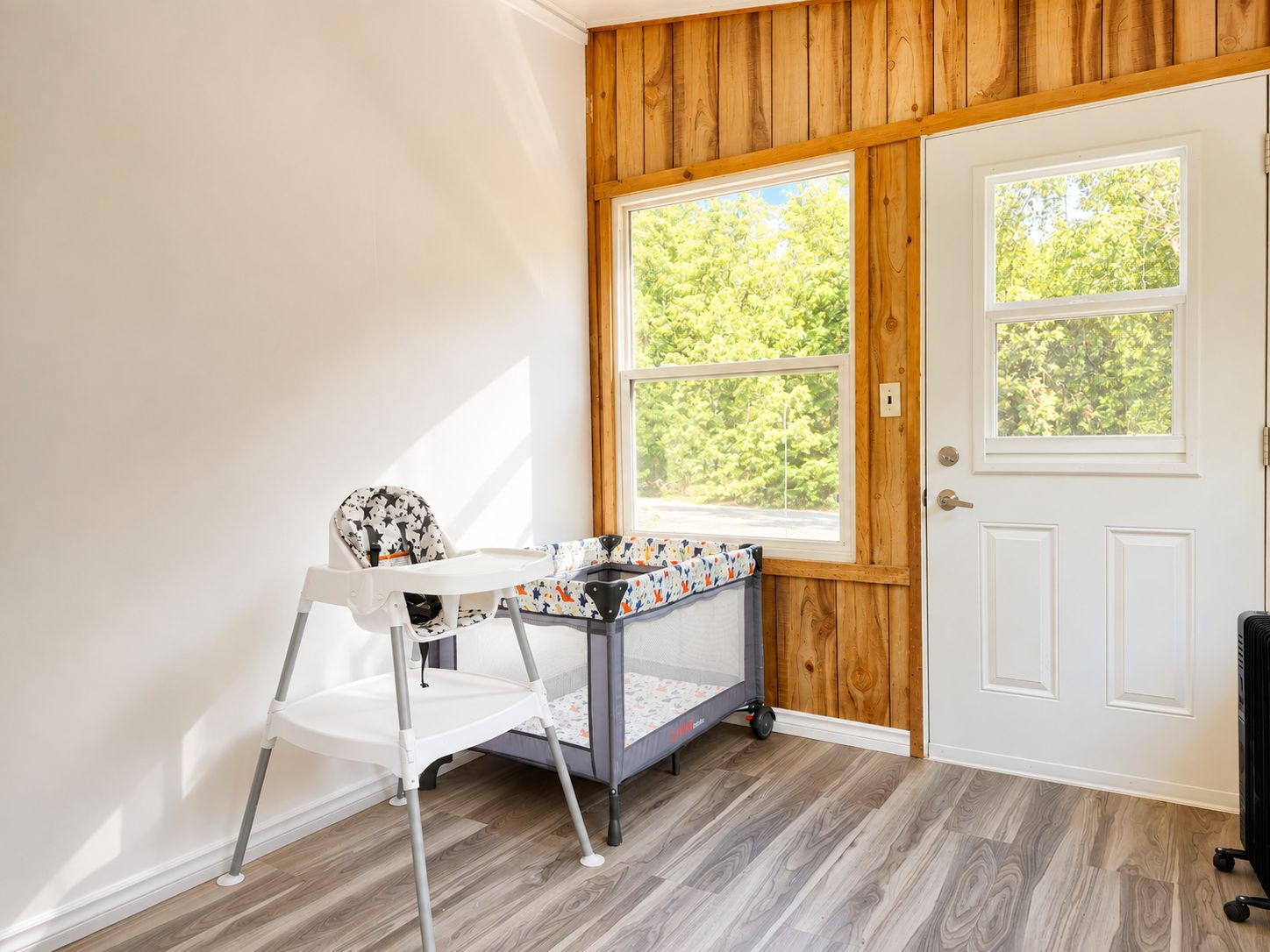 Une pièce avec une porte blanche, une fenêtre donnant sur des arbres, une chaise blanche et une petite table recouverte d’une nappe à motifs.