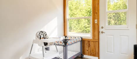 Una habitación con una puerta blanca, una ventana que da a los árboles, una silla blanca y una mesa pequeña con mantel estampado.