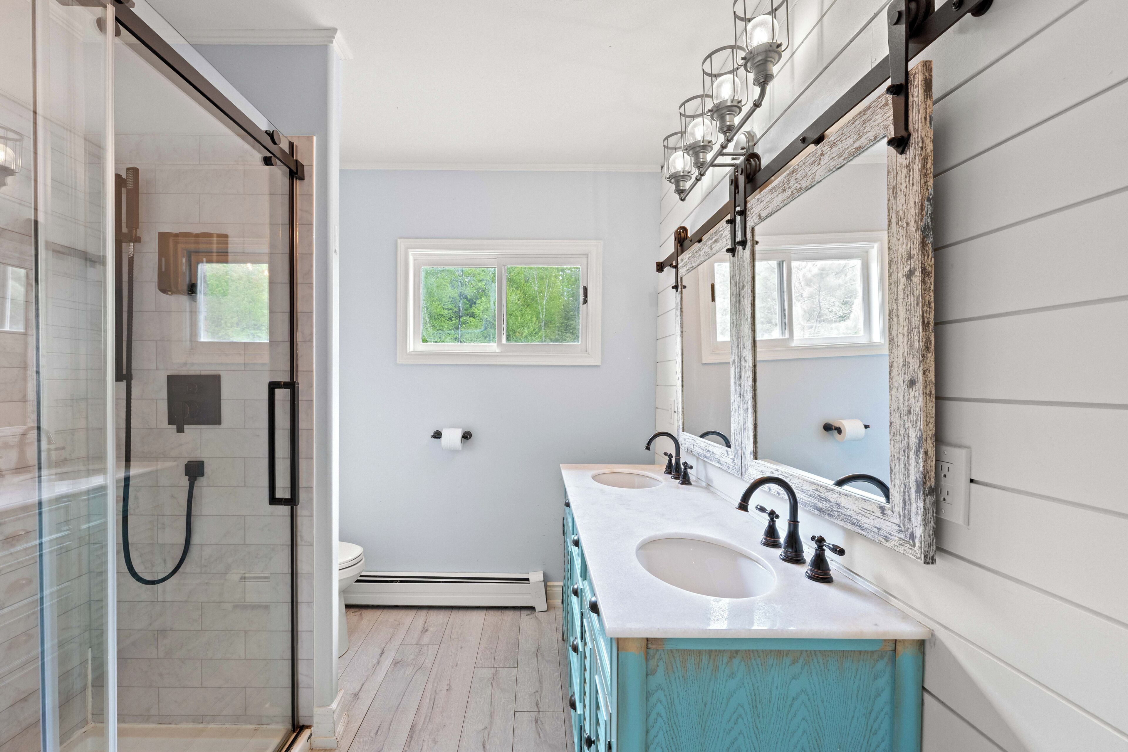 Un baño con cabina de ducha de vidrio, dos lavamanos con grifos de estilo vintage y un espejo grande con marco desgastado.