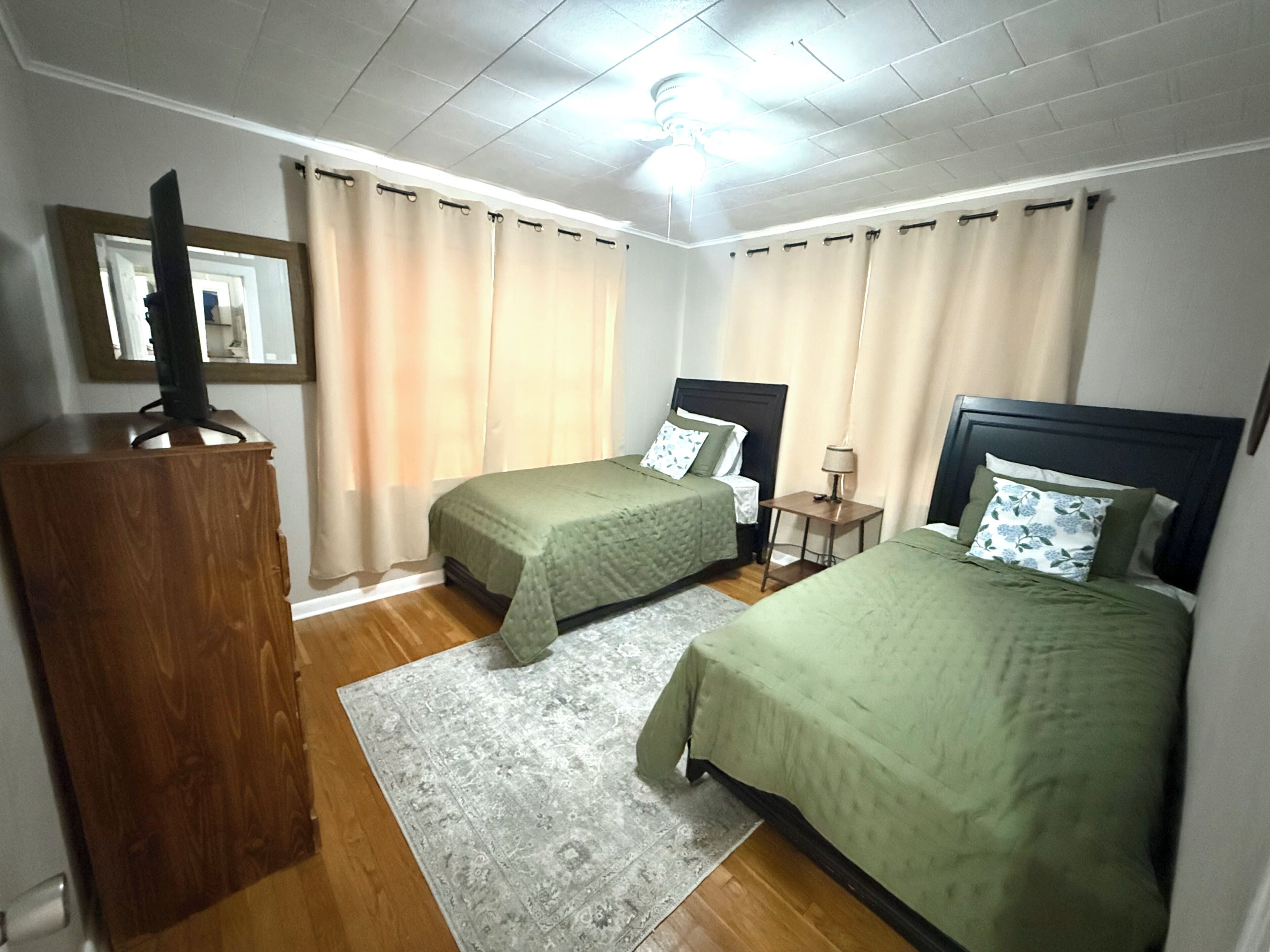 A hotel room with two single beds, a wooden desk, a television, and a ceiling fan.