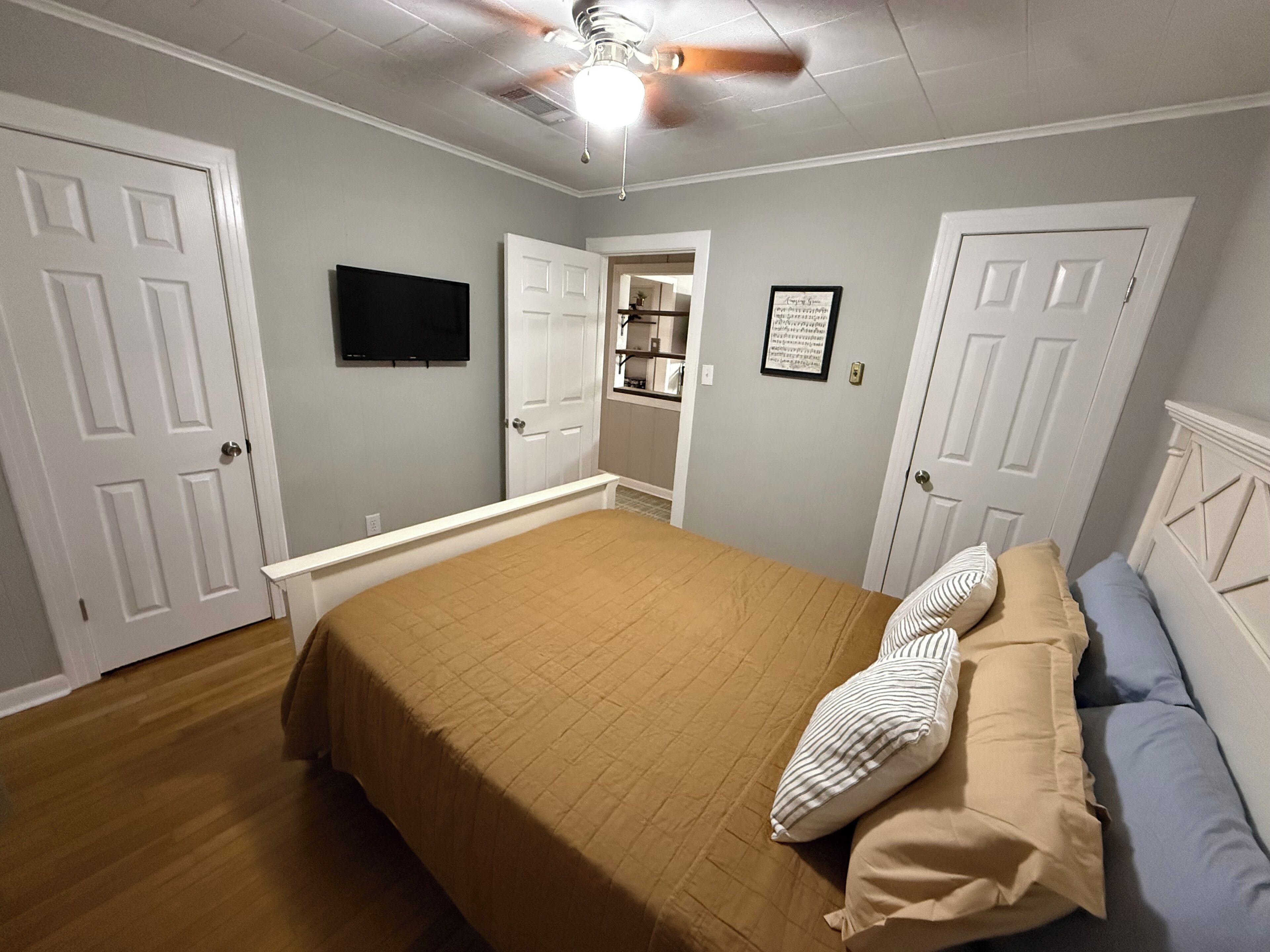 A bedroom with a bed, a ceiling fan, a television, and a door leading to a bathroom.