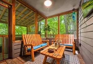 A wooden deck with two chairs and a table, surrounded by large windows with a forest view. - Rustic Cabin with Fire Pit and Hot Tub (Pigeon Forge)