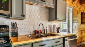 Una cocina con un lavamanos de acero inoxidable, una estufa y un lavavajillas. Se observan armarios con pomos, un salpicadero de azulejos hexagonales y un ventanal que proporciona luz natural.
